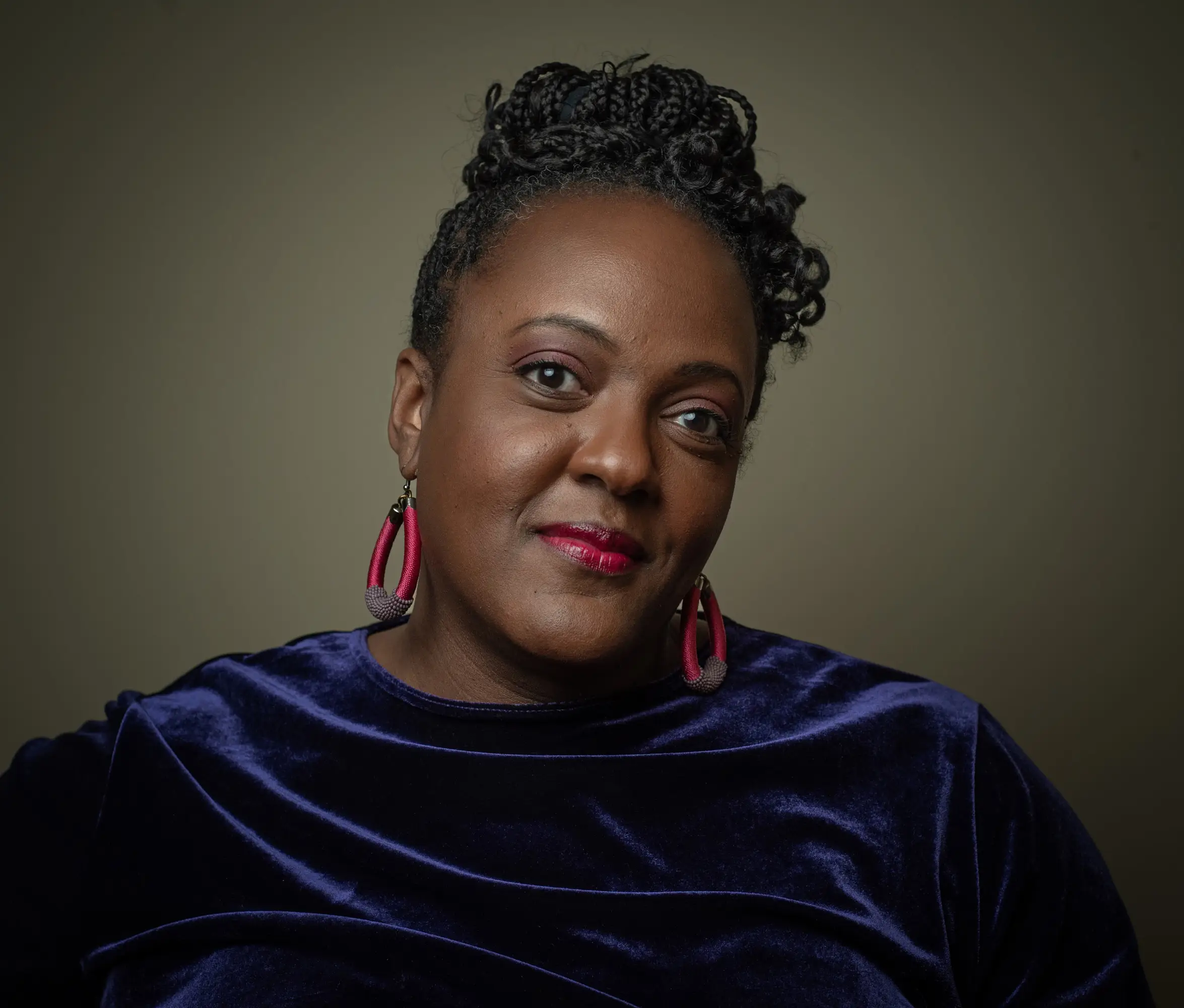 A Black woman with braided hair updo wearing a blue velvet top and pink hoop earrings poses against a dark backdrop.