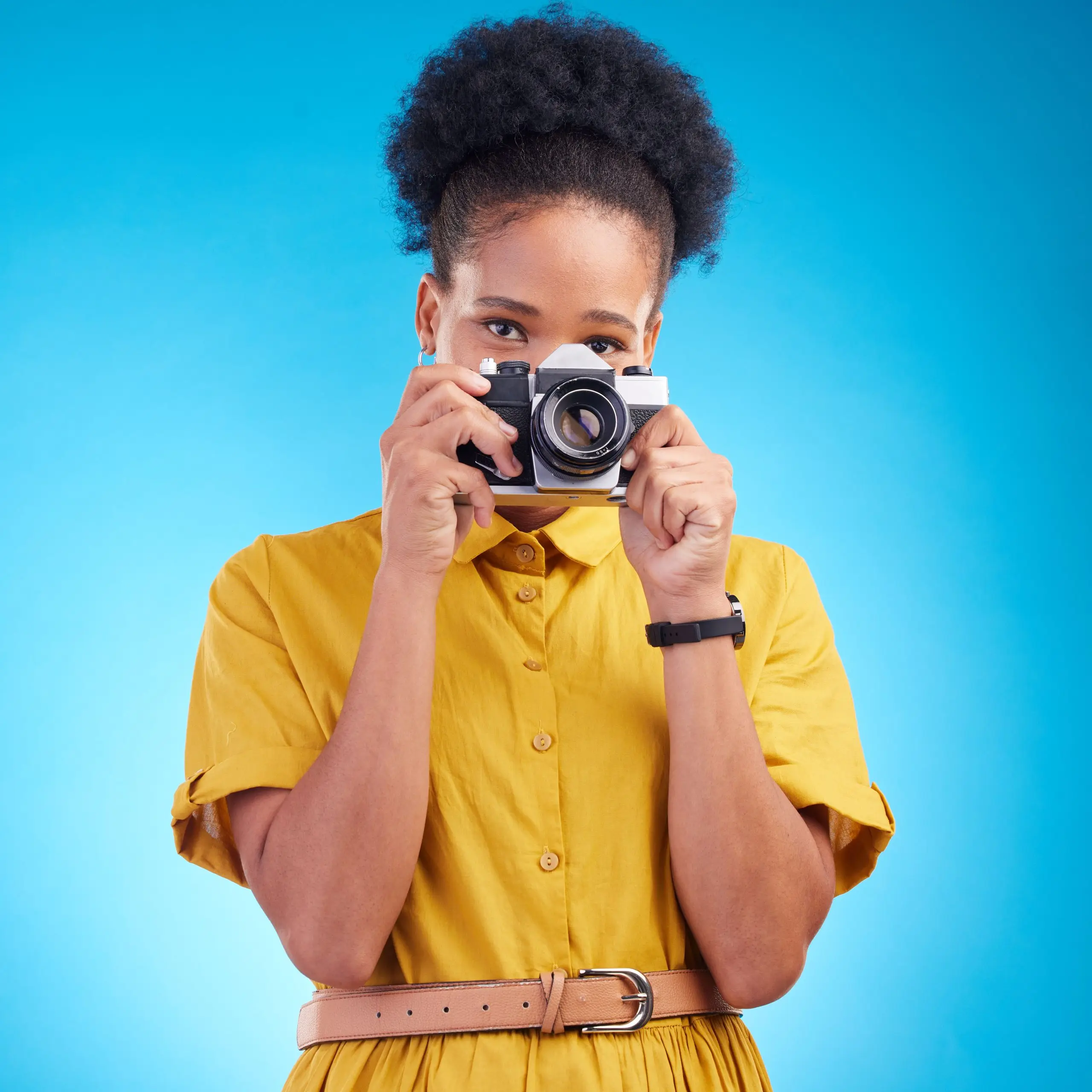 A confident Black woman photographer in a yellow dress holds a vintage camera to her face against a blue background.